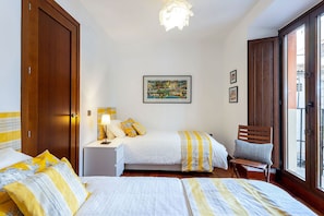 Un dormitorio con dos camas, una mesita de noche, una silla, un cuadro y una ventana.