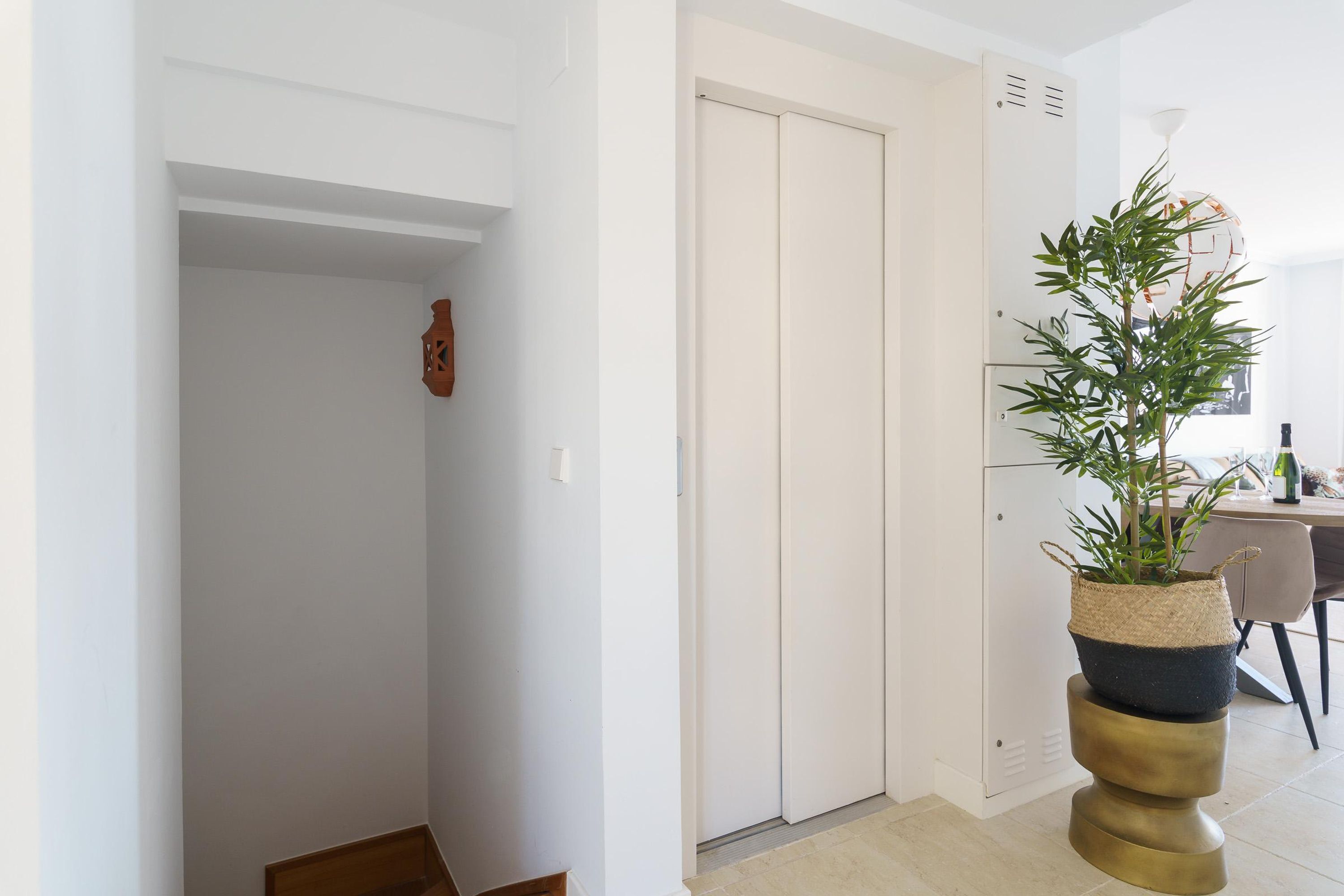 Un pasillo con una puerta blanca, una pequeña planta en una cesta tejida y un taburete de latón.