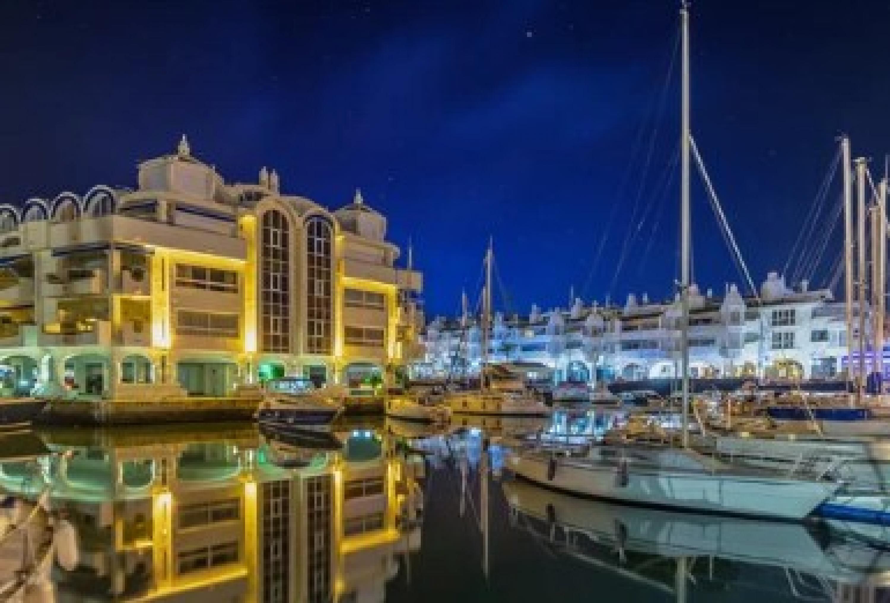 A marina with boats docked, a large building with a dome and arched windows, and a reflection in the water.