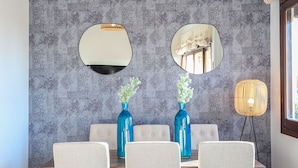 Een eethoek met behang met een patroon, een houten eettafel en stoelen. Er zijn twee wandspiegels en een hanglamp.
