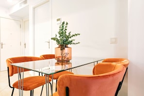 A modern dining area with a glass table and orange chairs, a potted plant, and white walls. - Luminous 1 Bedroom Apartment in the Heart of Seville Murillo V (Seville)