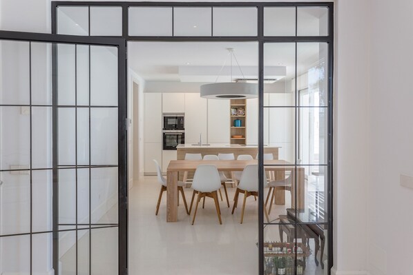 Un espace repas moderne avec une table en bois et des chaises blanches, une cuisine en arrière-plan et une grande fenêtre offrant une belle vue.