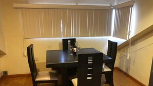 Een eetgedeelte met een donker houten tafel en stoelen, een klein bijzettafeltje met een fles en glazen, en een raam met jaloezieën.