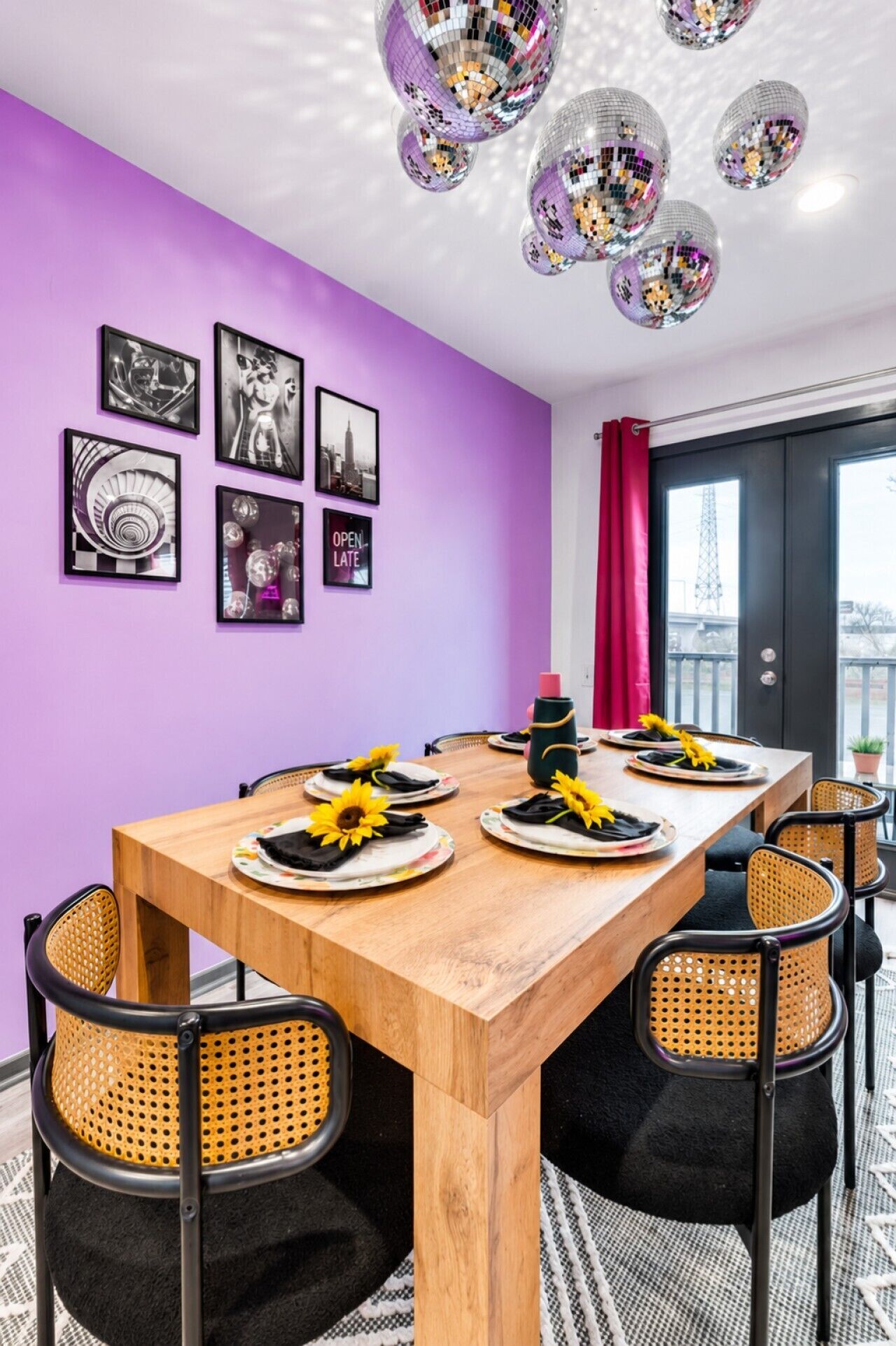 A dining area with a wooden table set for six, surrounded by wicker chairs, with a purple wall and ceiling disco balls.
