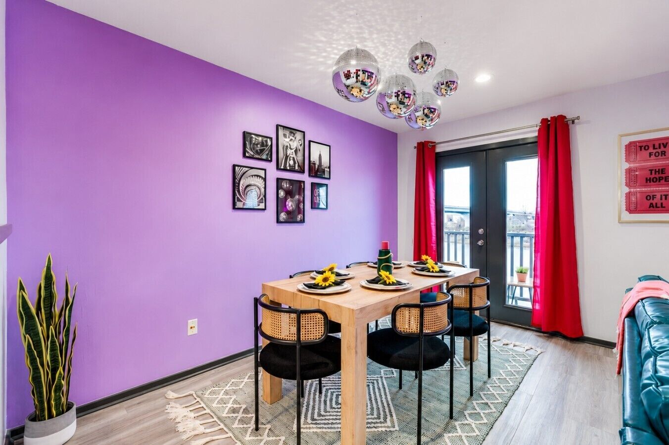 A dining area with a wooden table set for four, surrounded by chairs, with a purple wall and a series of framed pictures above.