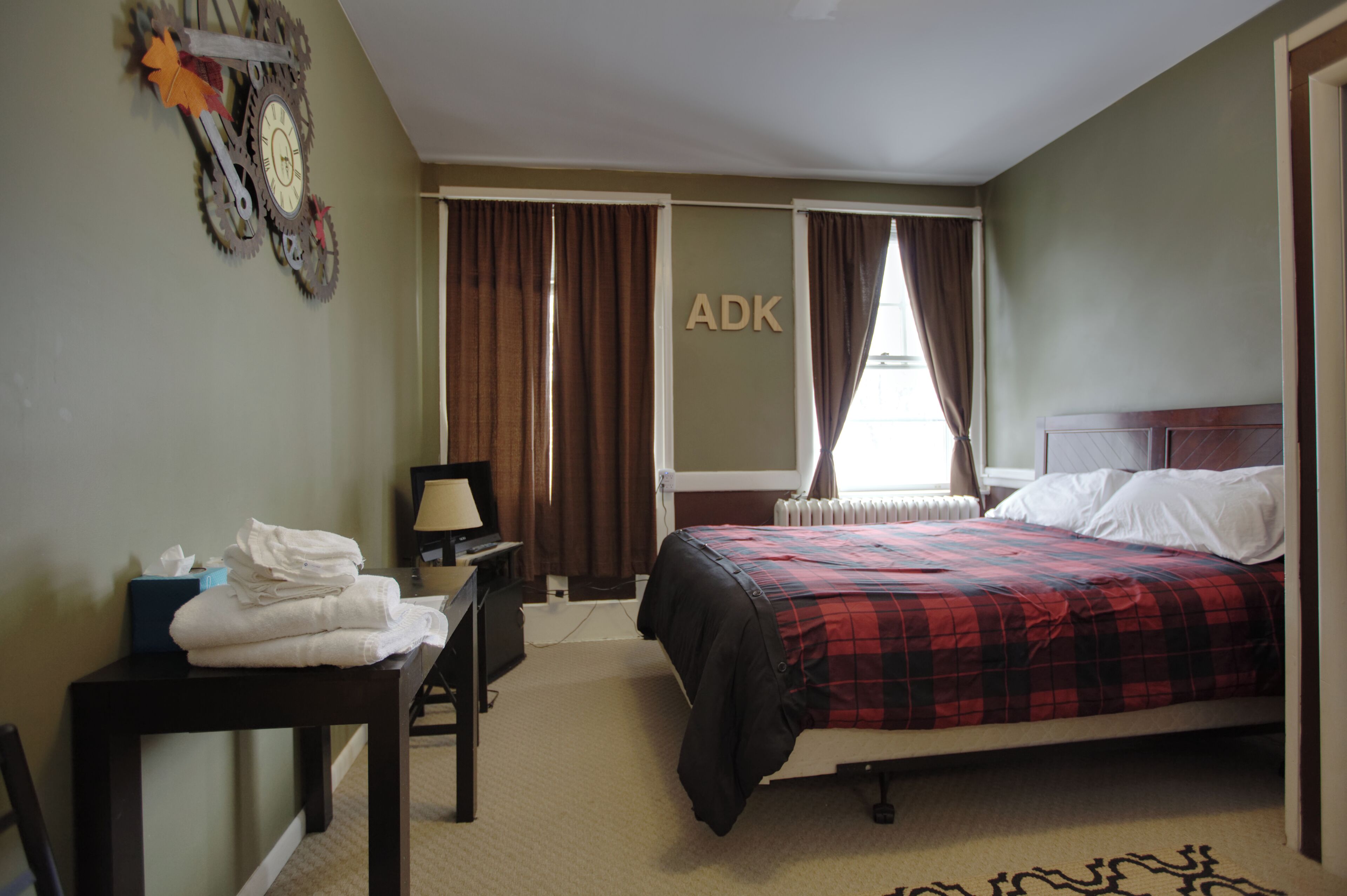 A hotel room with a bed, a desk with a lamp, a radiator, and a wall-mounted clock.