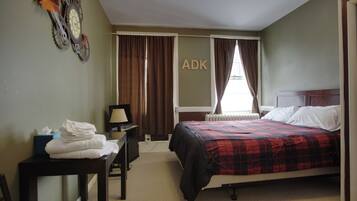 A hotel room with a bed, a desk with a lamp, a radiator, and a wall-mounted clock.