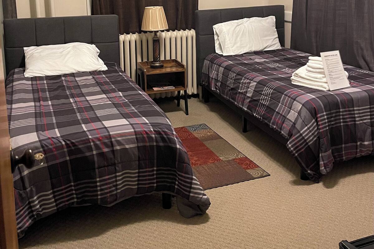 A hotel room with two beds, a nightstand with a lamp, a radiator, and a small shelf.