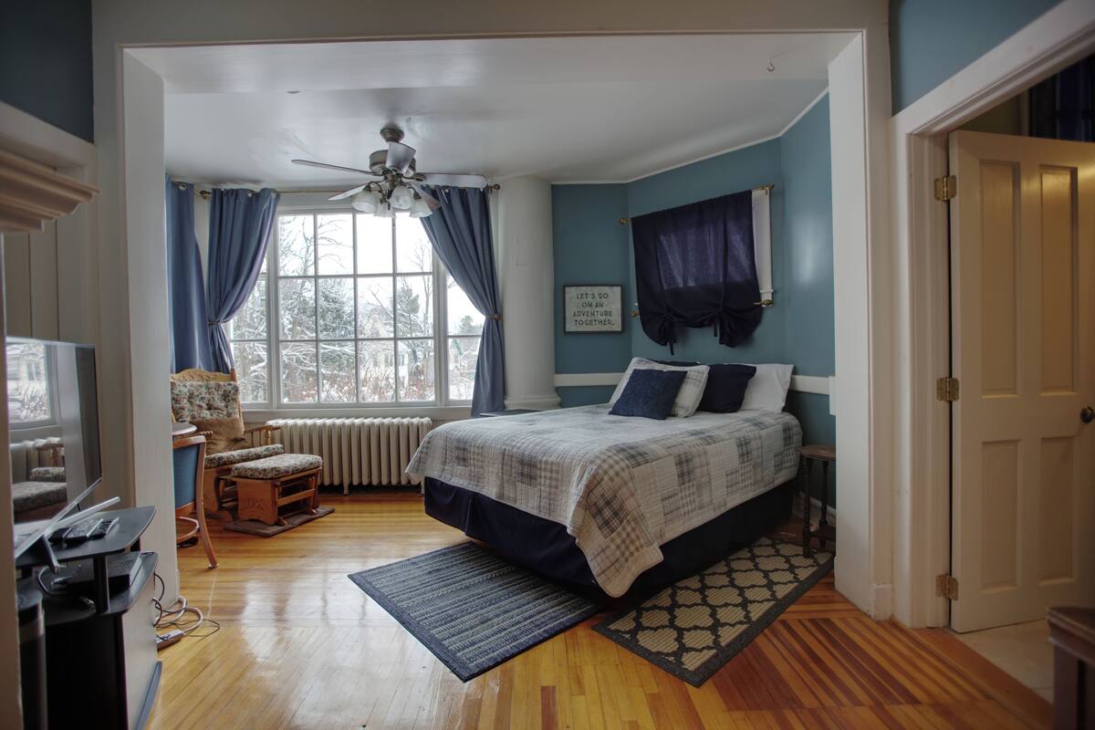 A bedroom with a bed, a desk, a chair, a window with curtains, and a ceiling fan.