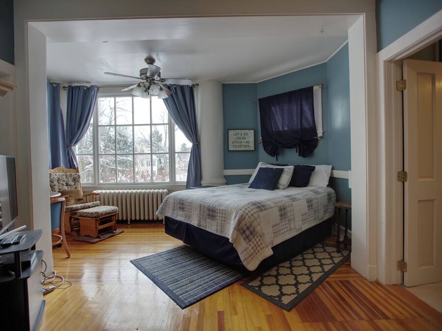 A bedroom with a bed, a desk, a chair, a window with curtains, and a ceiling fan.