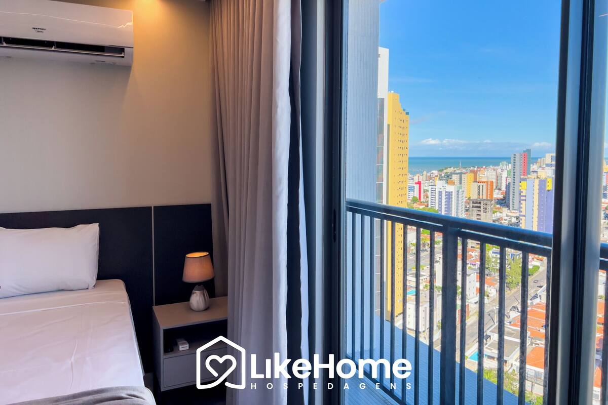 A hotel room with a bed, bedside lamp, air conditioning unit, and a view of a cityscape through large windows.