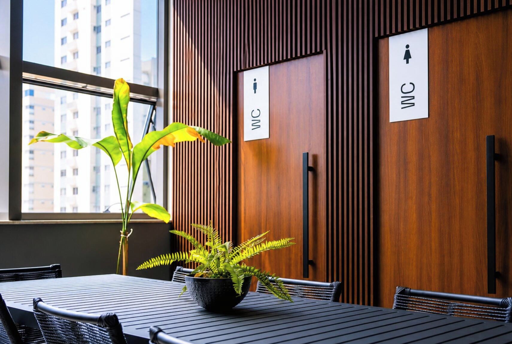 Interior moderno com parede de madeira, uma janela e uma mesa com cadeiras. Há placas indicando banheiros masculino e feminino.