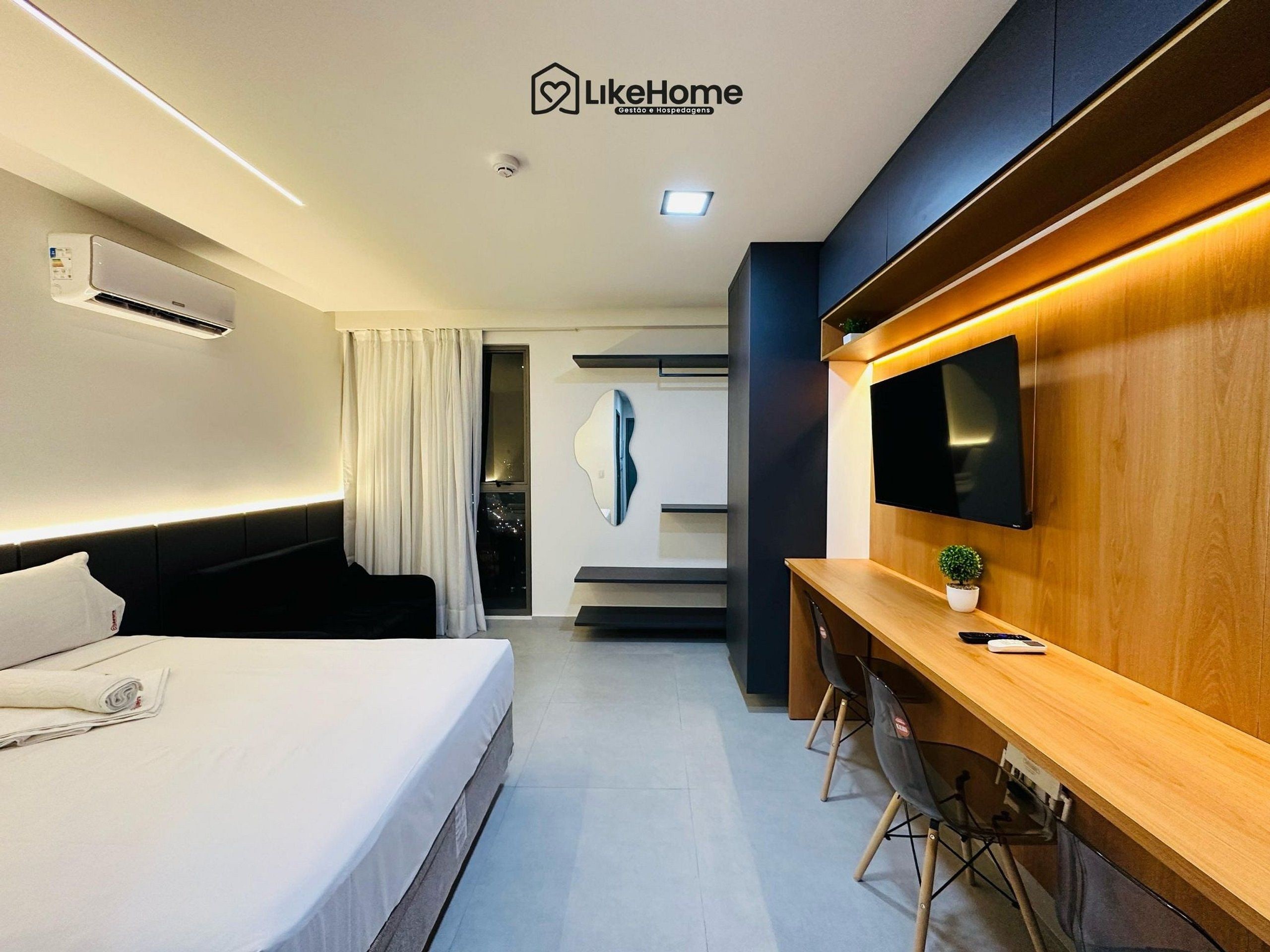 A modern hotel room with a bed, a desk with chairs, a flat-screen TV, and a wall-mounted air conditioner.