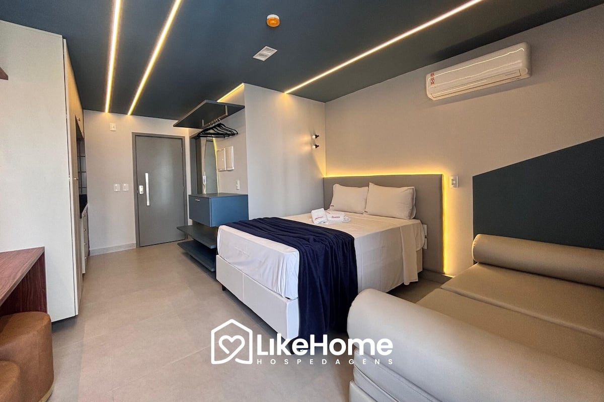 A modern hotel room with a bed, a sofa, a desk, and a mirror. There is an air conditioning unit on the wall.