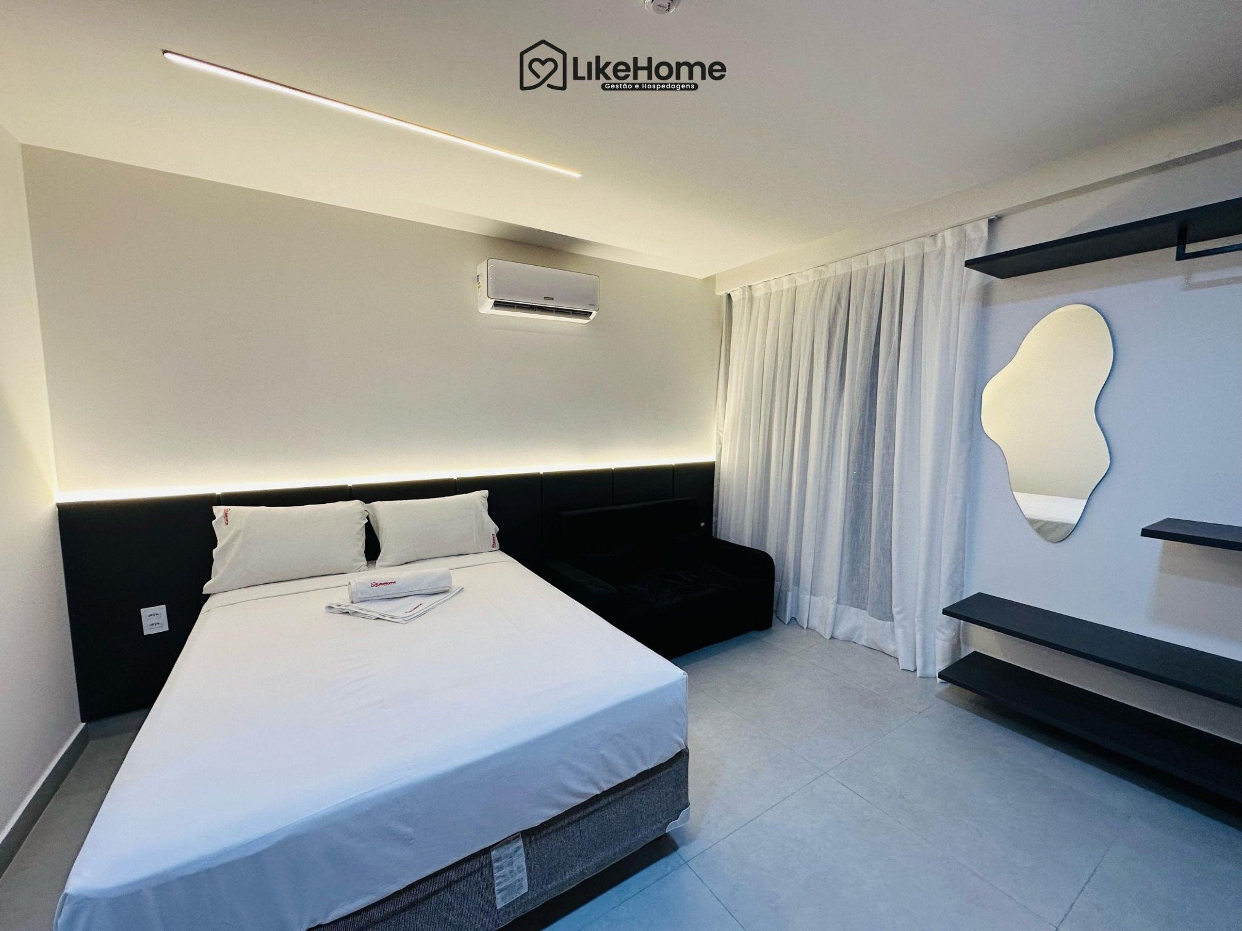 A modern hotel room with a bed, bedside table, air conditioning unit, and a wall-mounted mirror.