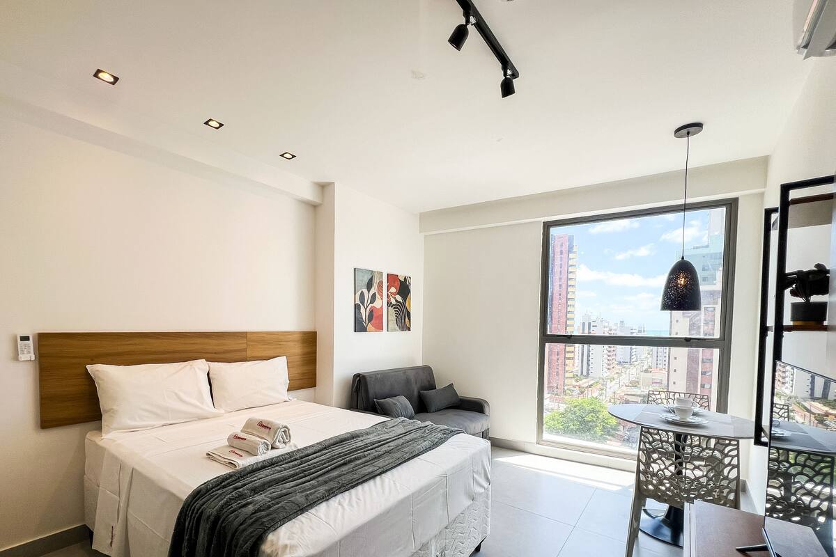 A modern hotel room with a large bed, a grey armchair, a small table with a cup, and a city view through a large window.