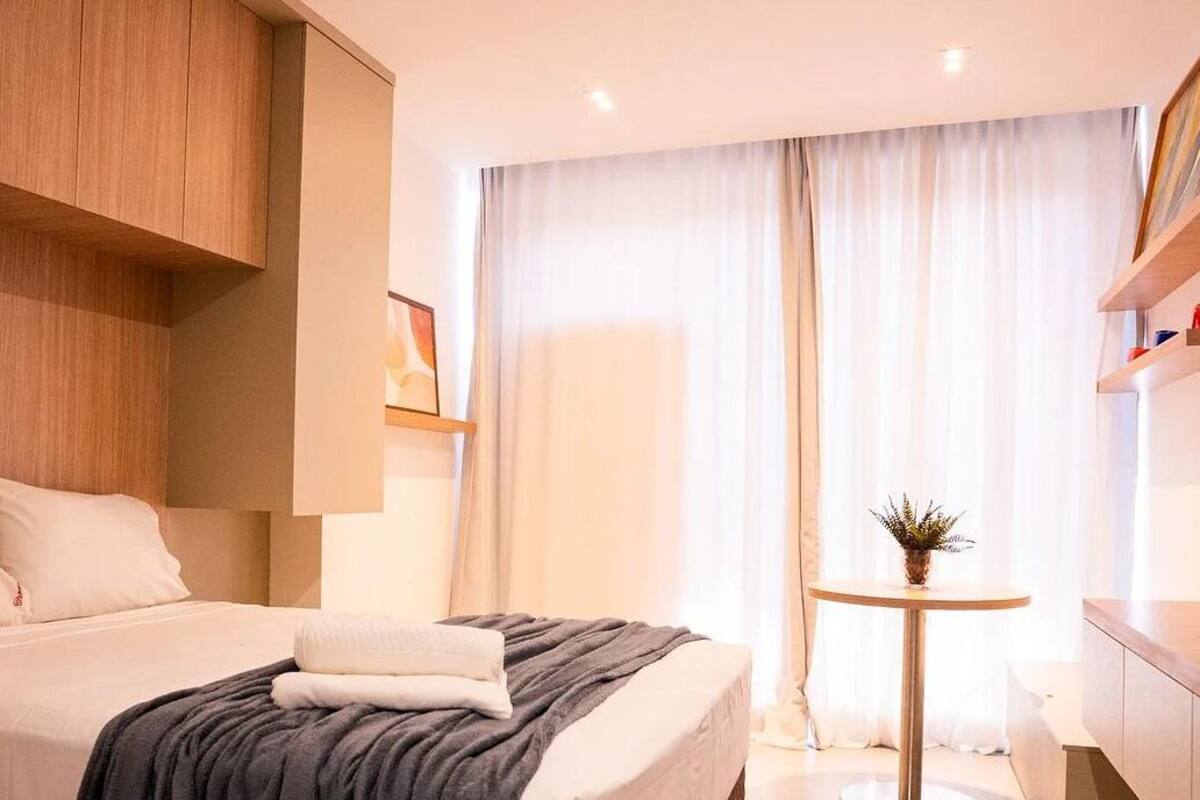 A modern hotel room with a large bed, a small round table, and a view of the city through sheer curtains.