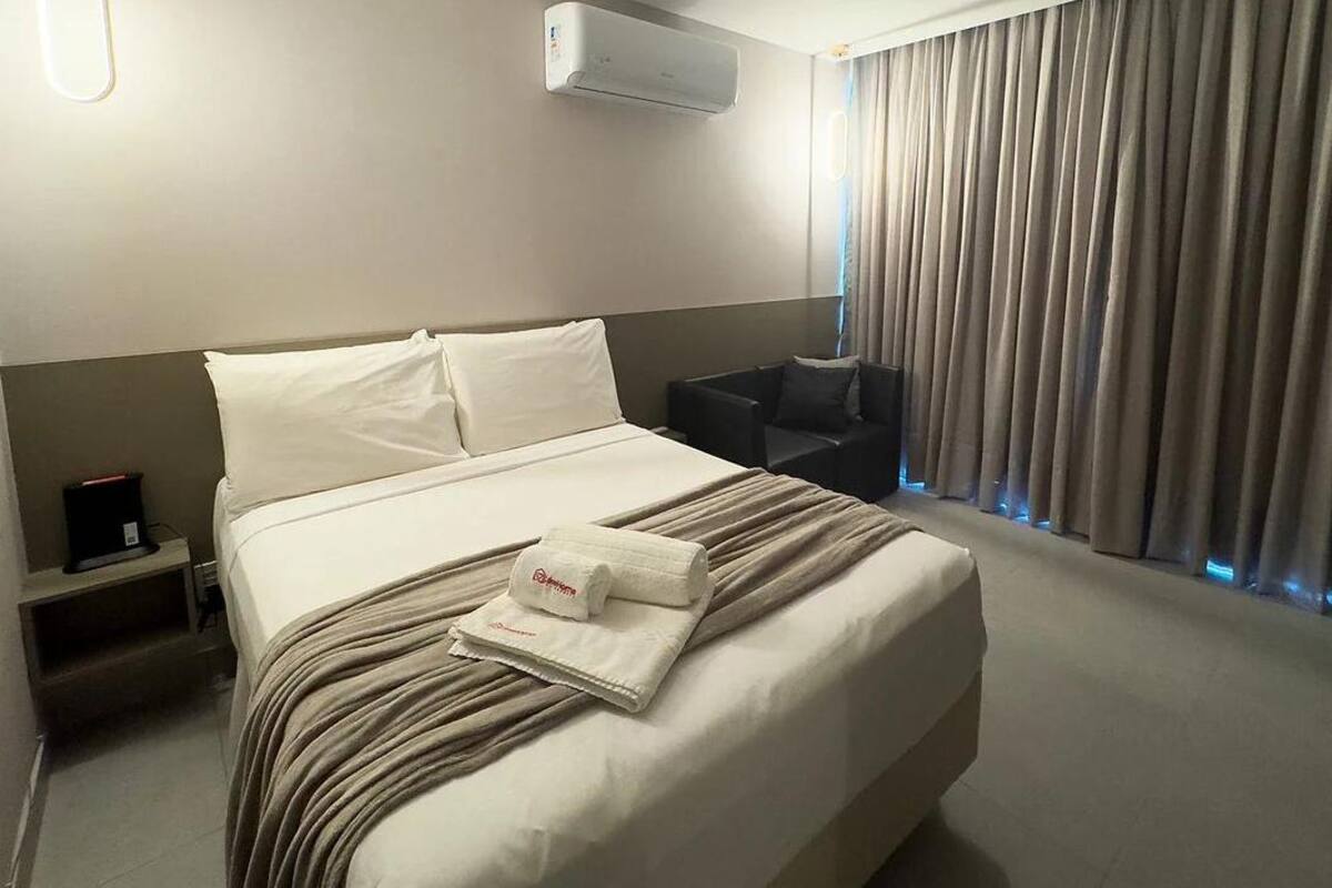A hotel room with a bed, a chair, a nightstand, and an air conditioning unit.
