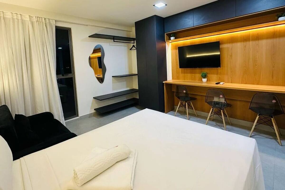 A modern hotel room with a large bed, a desk with two chairs, a TV mounted on a wooden wall, and a small plant on the desk.