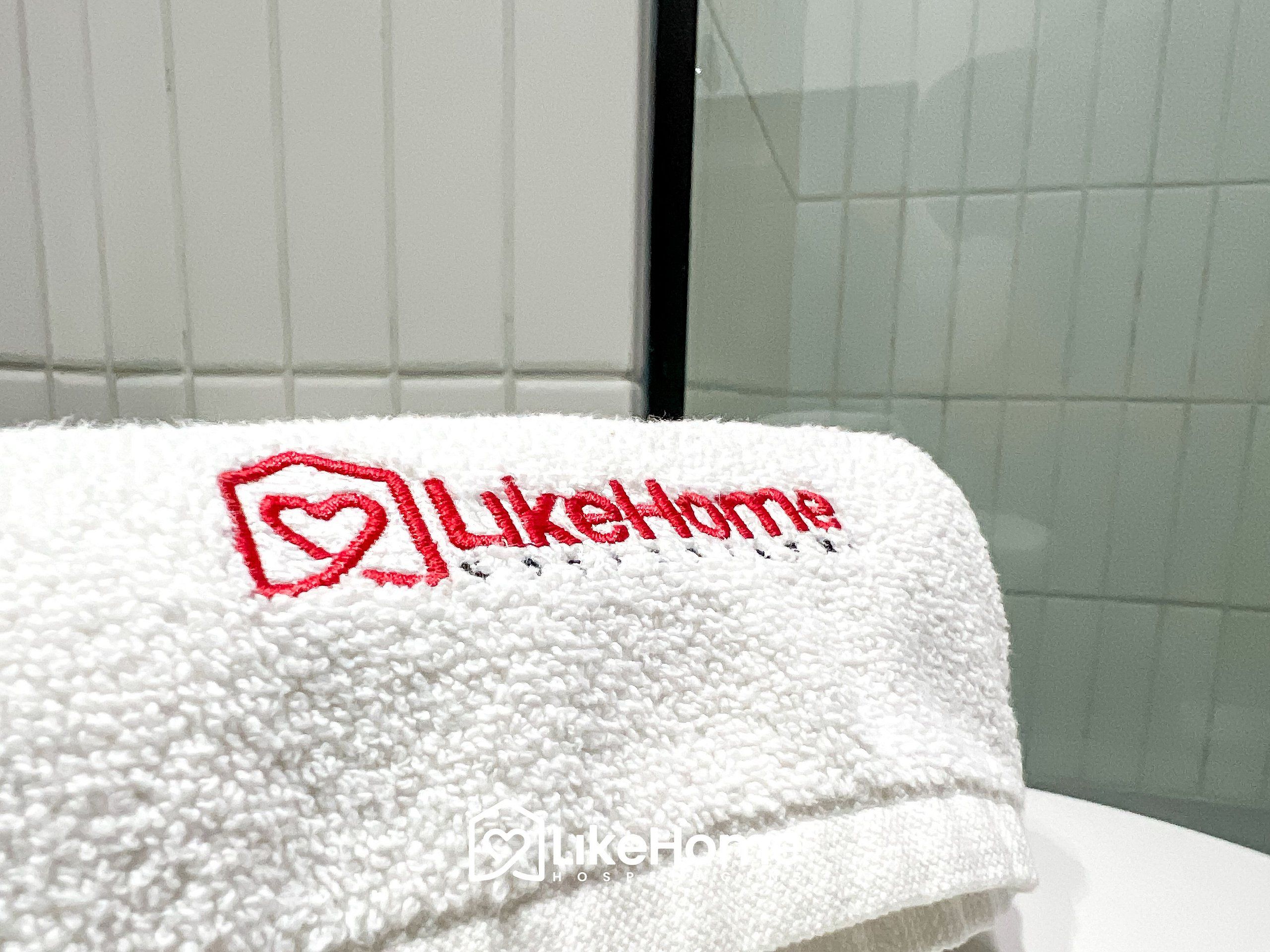 A white towel with a heart-shaped logo and the text 'likeHome' embroidered on it.