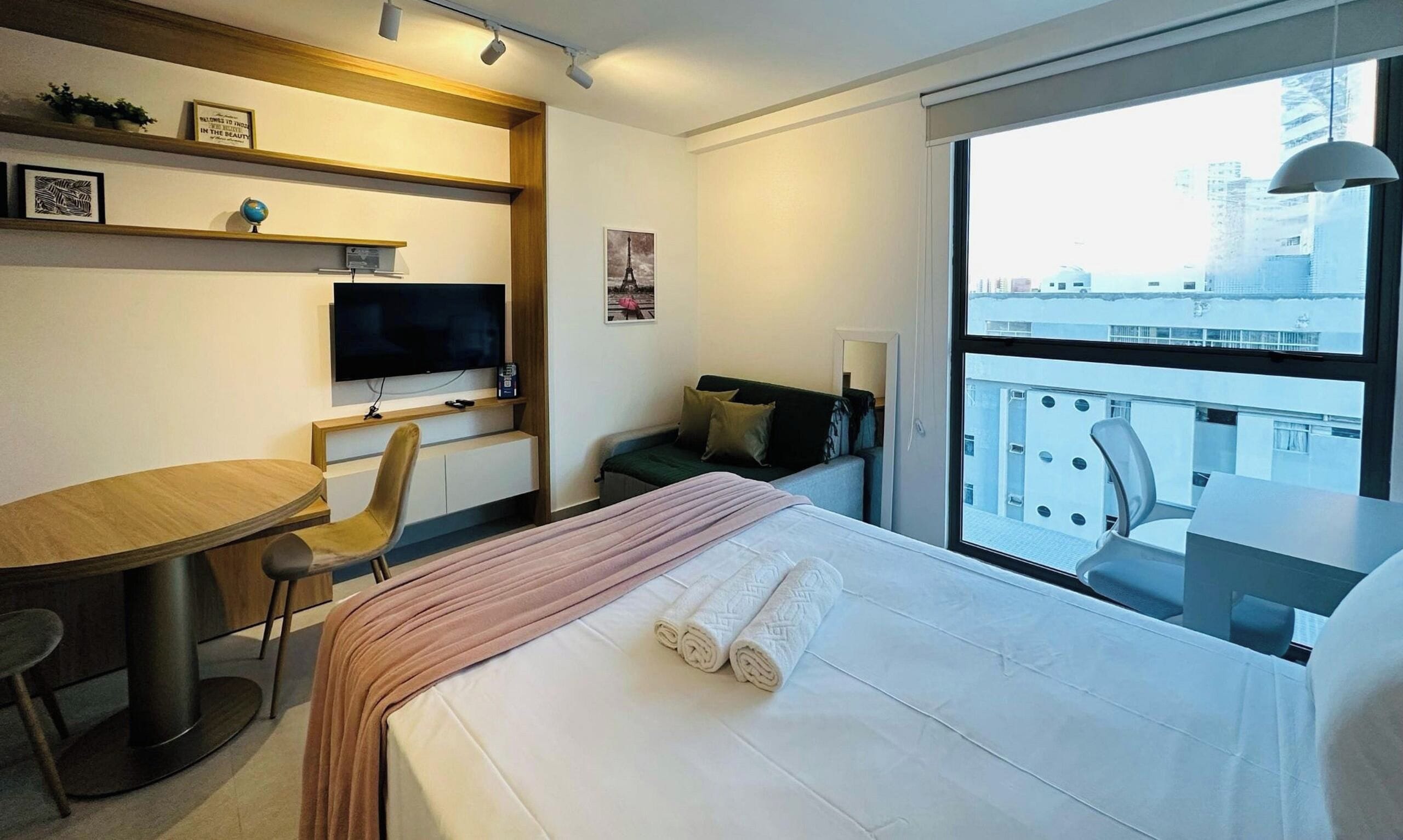 A hotel room with a bed, a desk, a chair, a television, and a large window offering a city view.