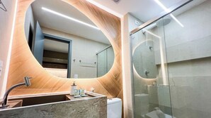 A modern bathroom with a curved mirror, a marble sink, and a glass shower enclosure. - Simple Smart - LikeHome Hospedagens (João Pessoa)