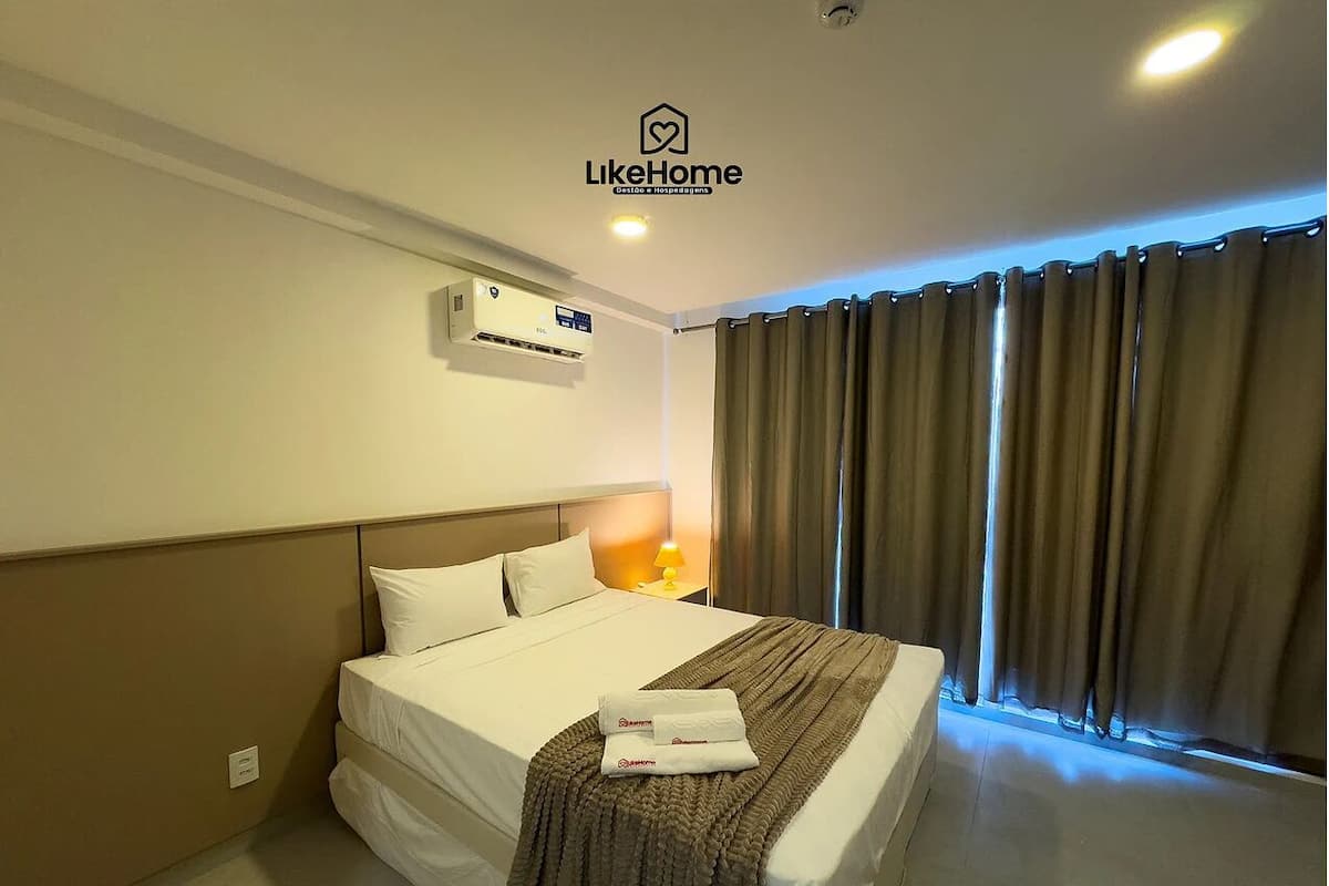A hotel room with a bed, a bedside table with a lamp, a wall-mounted air conditioner, and a window with curtains.