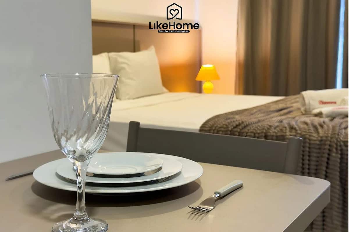 A hotel room with a neatly made bed, a bedside lamp, and a dining area with a glass, plates, and cutlery.