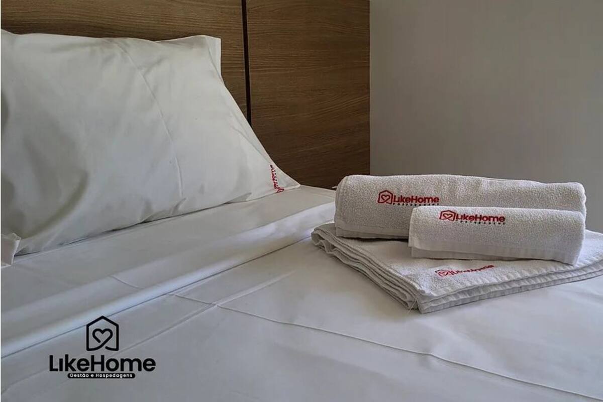 A neatly made bed with white linens and two towels with the LikeHome logo.