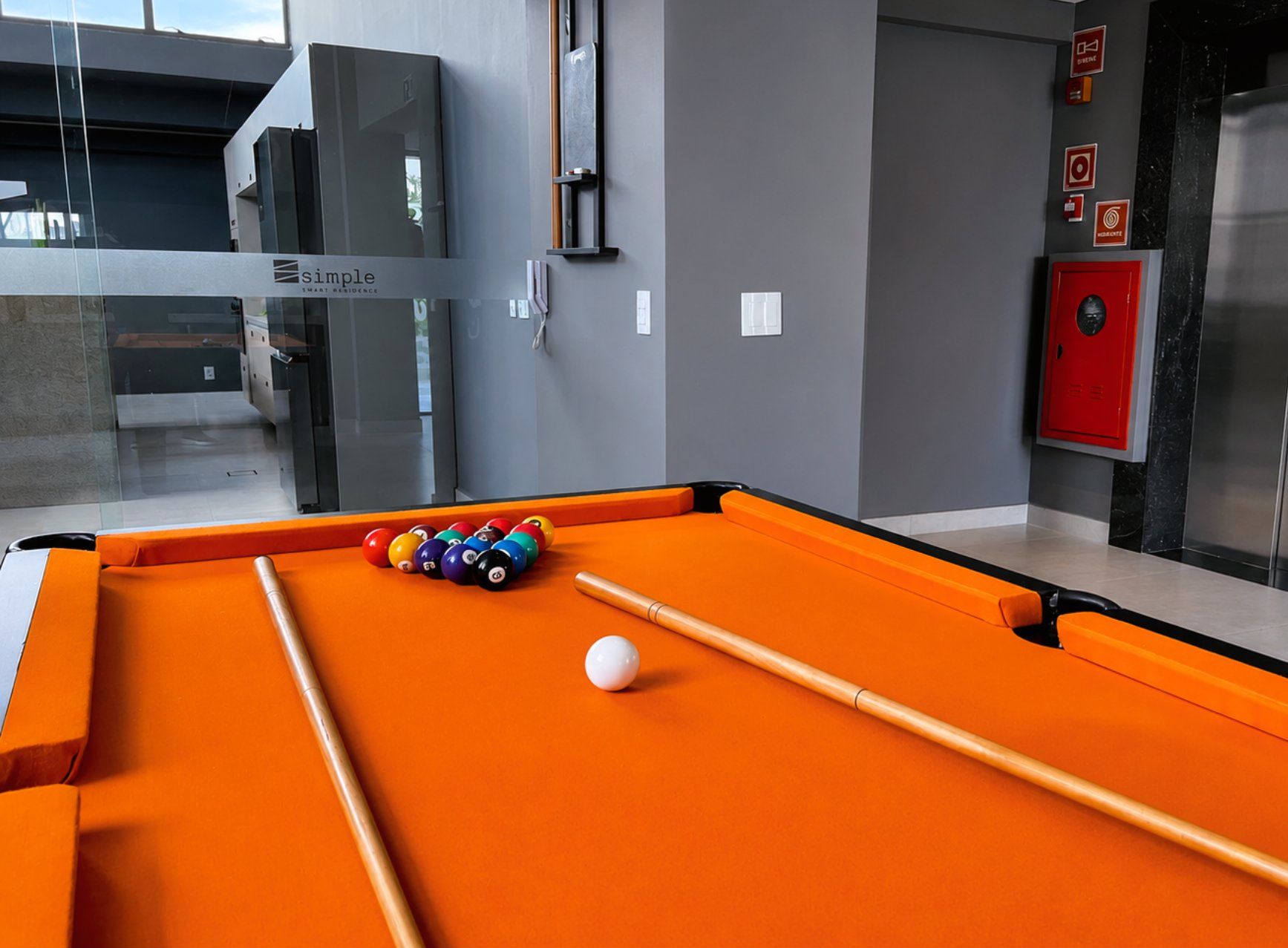 A pool table with balls and a cue stick, a red door, and a glass partition with signage.