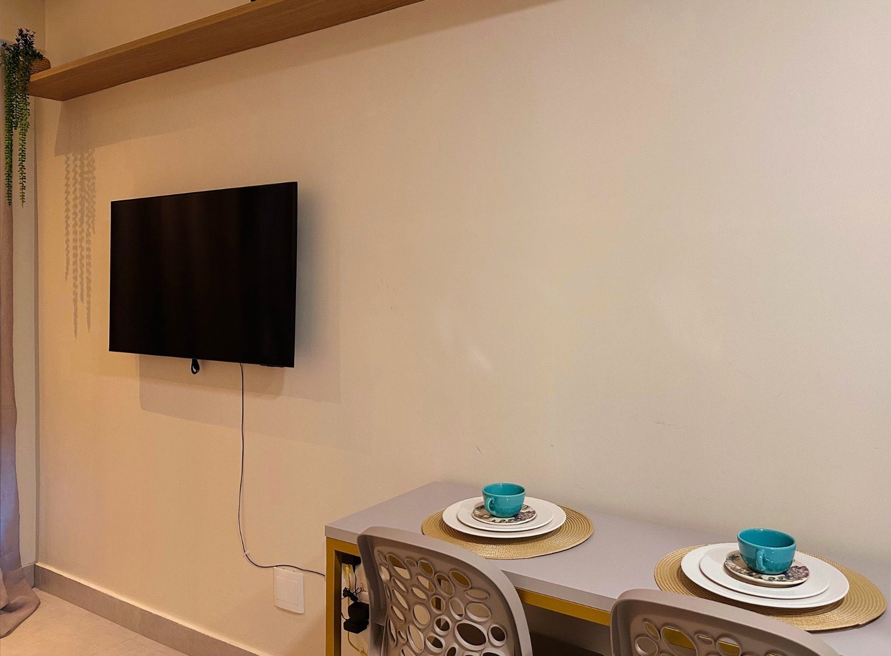 A room with a flat-screen TV mounted on the wall, a dining table with two chairs, and a small plant on the left.