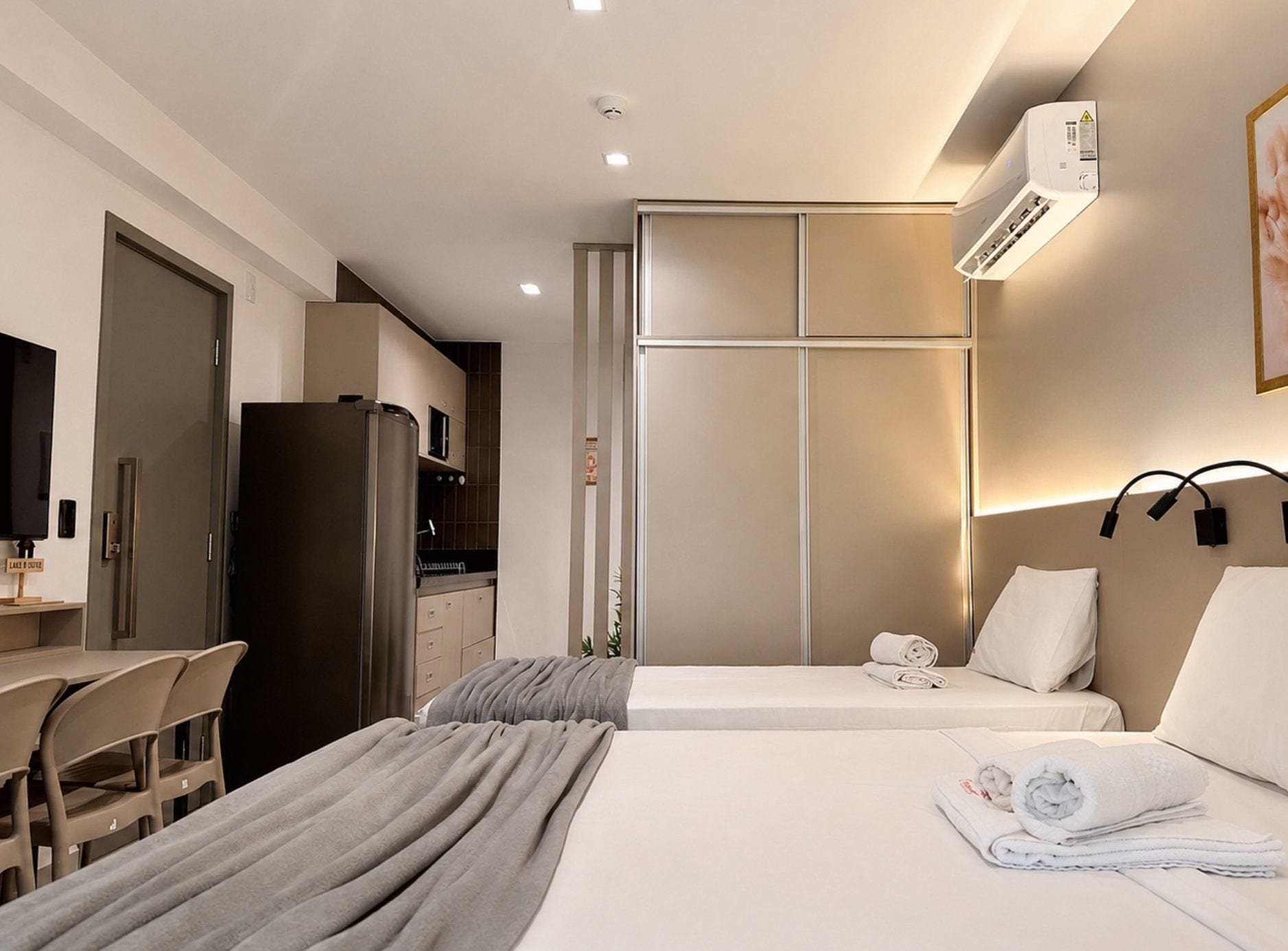 A hotel room with a bed, a desk, a chair, a kitchenette, and an air conditioning unit.