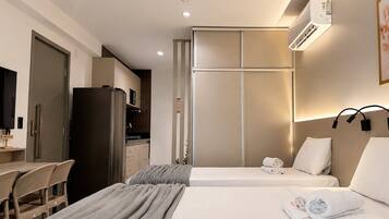 A hotel room with a bed, a desk, a chair, a kitchenette, and an air conditioning unit.