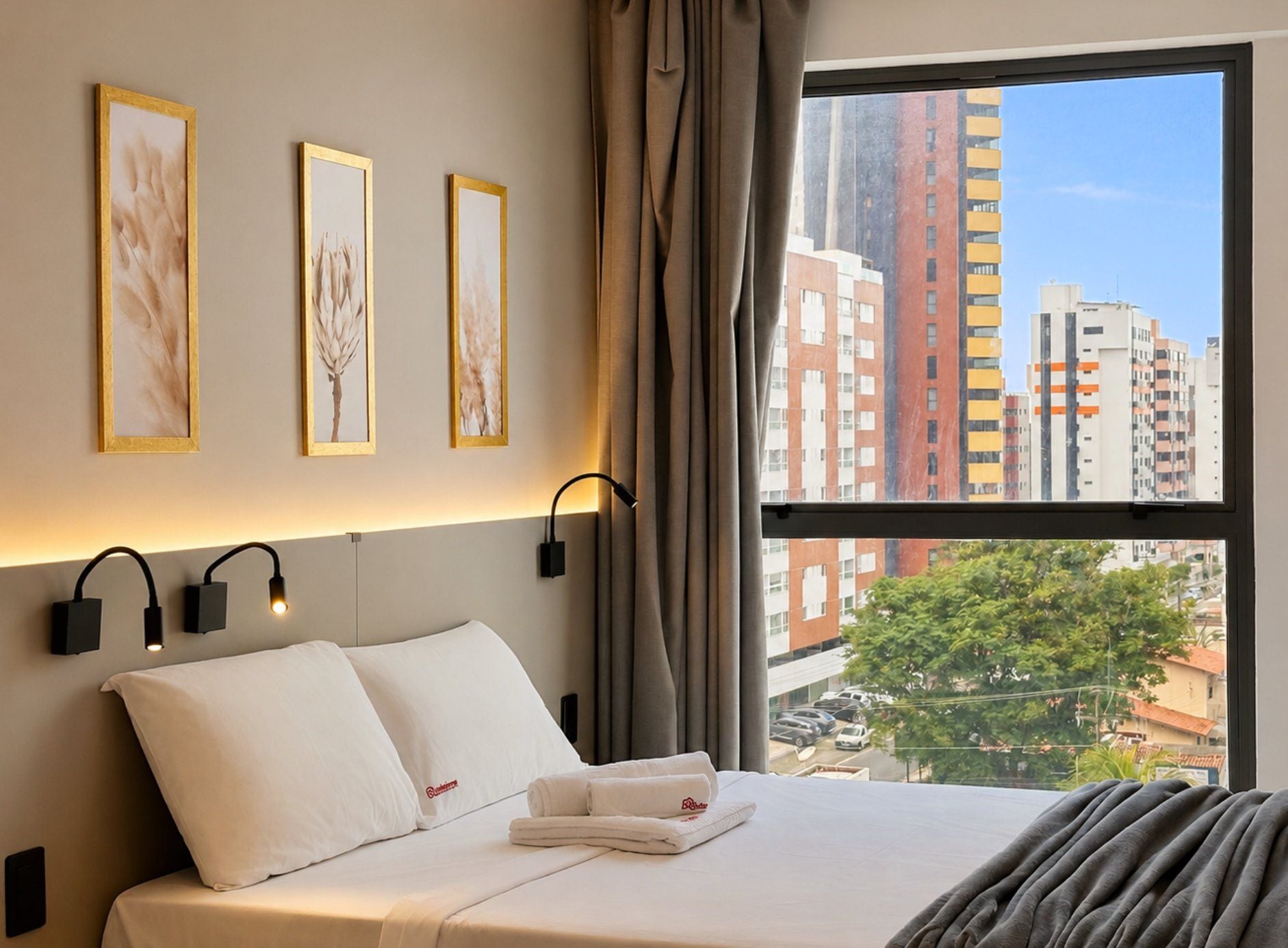 Een hotelkamer met een bed, witte kussens, een raam met gordijnen en wandlampen.