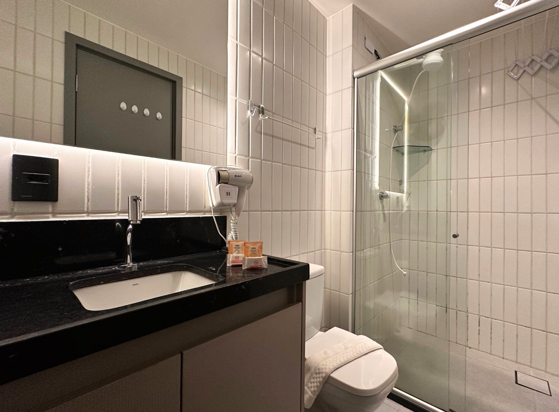 Un baño moderno con encimera negra, lavabo blanco, secador de pelo y cabina de ducha de vidrio.