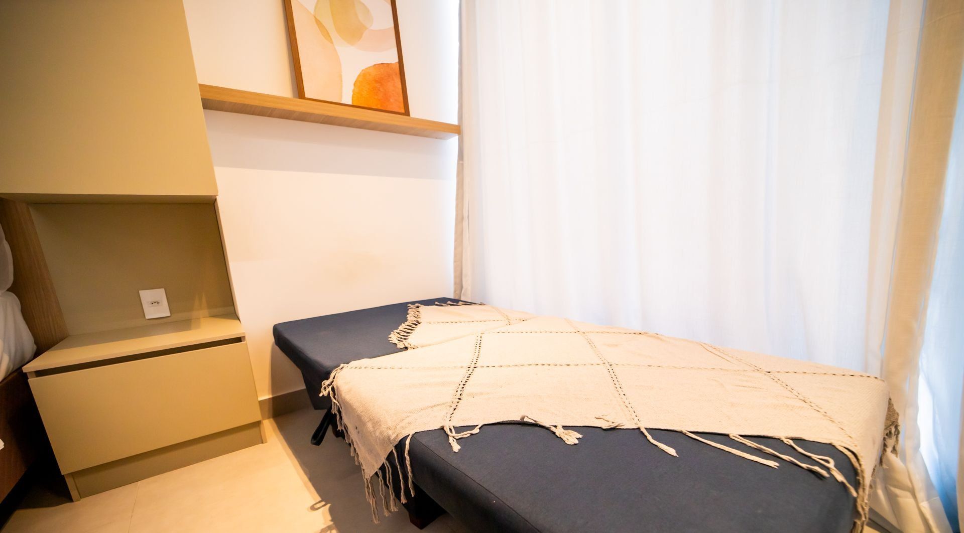 Quarto pequeno e reservado, com uma cama de solteiro, mesa de cabeceira e prateleira na parede.