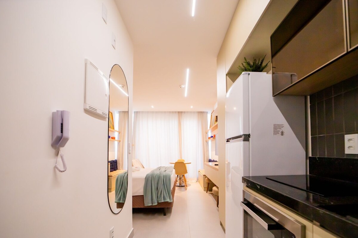 Une chambre d’hôtel compacte comprenant un coin cuisine, un lit avec une couverture bleue, un petit coin salon et un miroir.