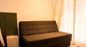 A small, dark-colored sofa in a corner of a room with a white wall and a window with sheer curtains. - Simple Smart - LikeHome Hospedagens (João Pessoa)