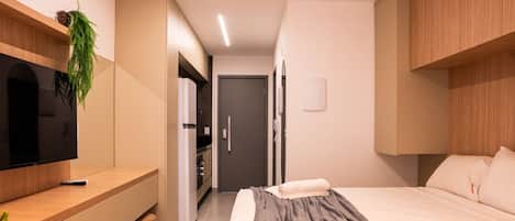 A compact hotel room with a bed, bedside table, TV, and a kitchenette area.