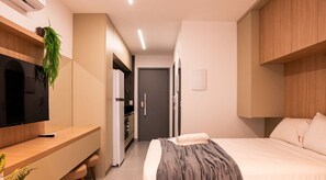 A compact hotel room with a bed, bedside table, TV, and a kitchenette area. - Simple Smart - LikeHome Hospedagens (João Pessoa)