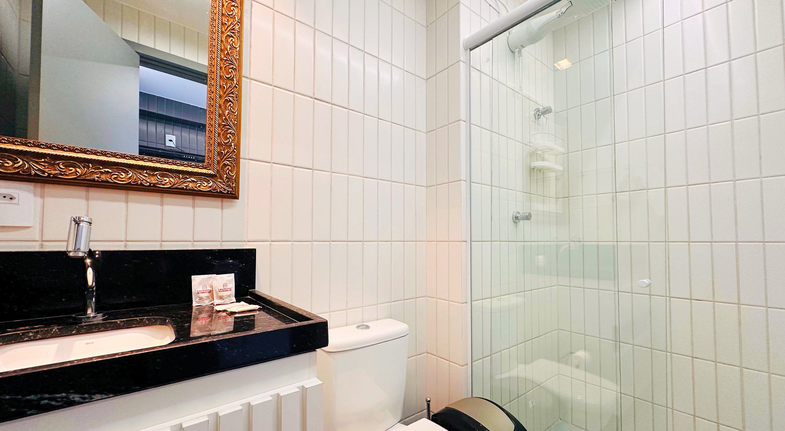 Un baño con encimera negra, baldosas blancas, cabina de ducha de cristal y espejo con marco dorado.