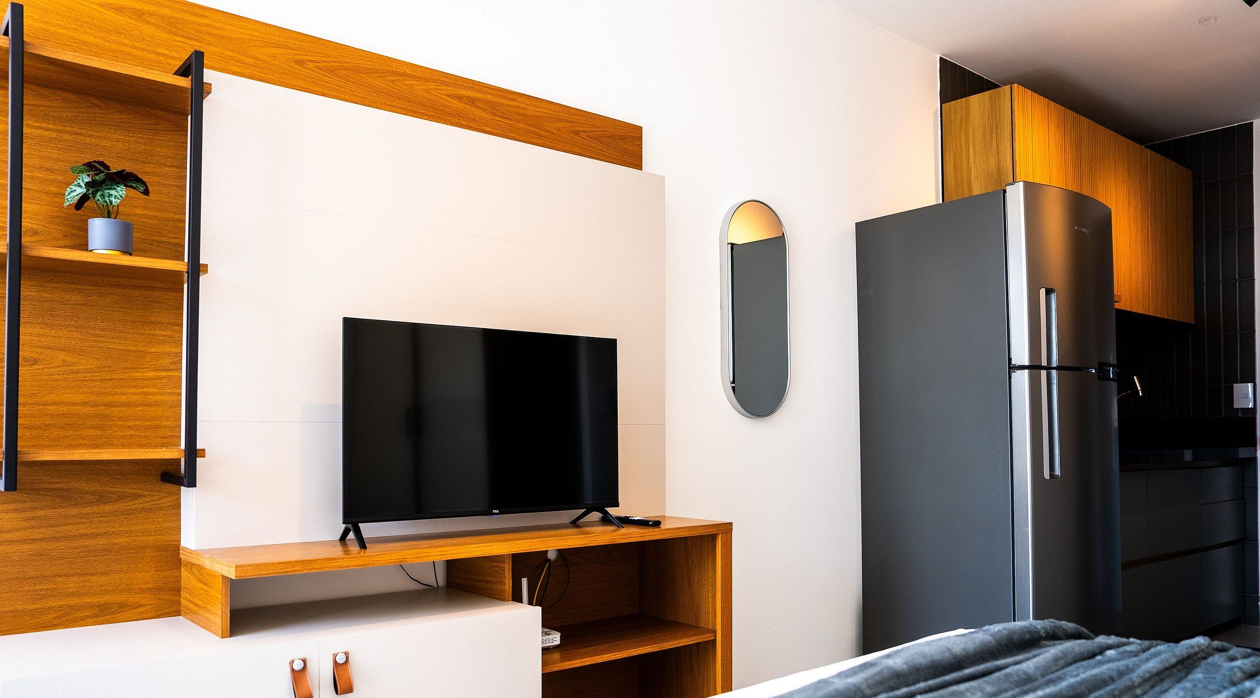 Una habitación moderna con una televisión de pantalla plana sobre un soporte de madera, una pequeña planta en maceta, un espejo y un refrigerador empotrado.