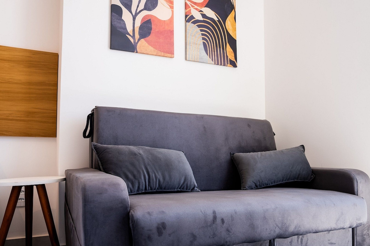 Ein graues Sofa mit zwei Kissen, ein weißer Tisch mit Holzfuß und abstrakte Kunst am Wand.