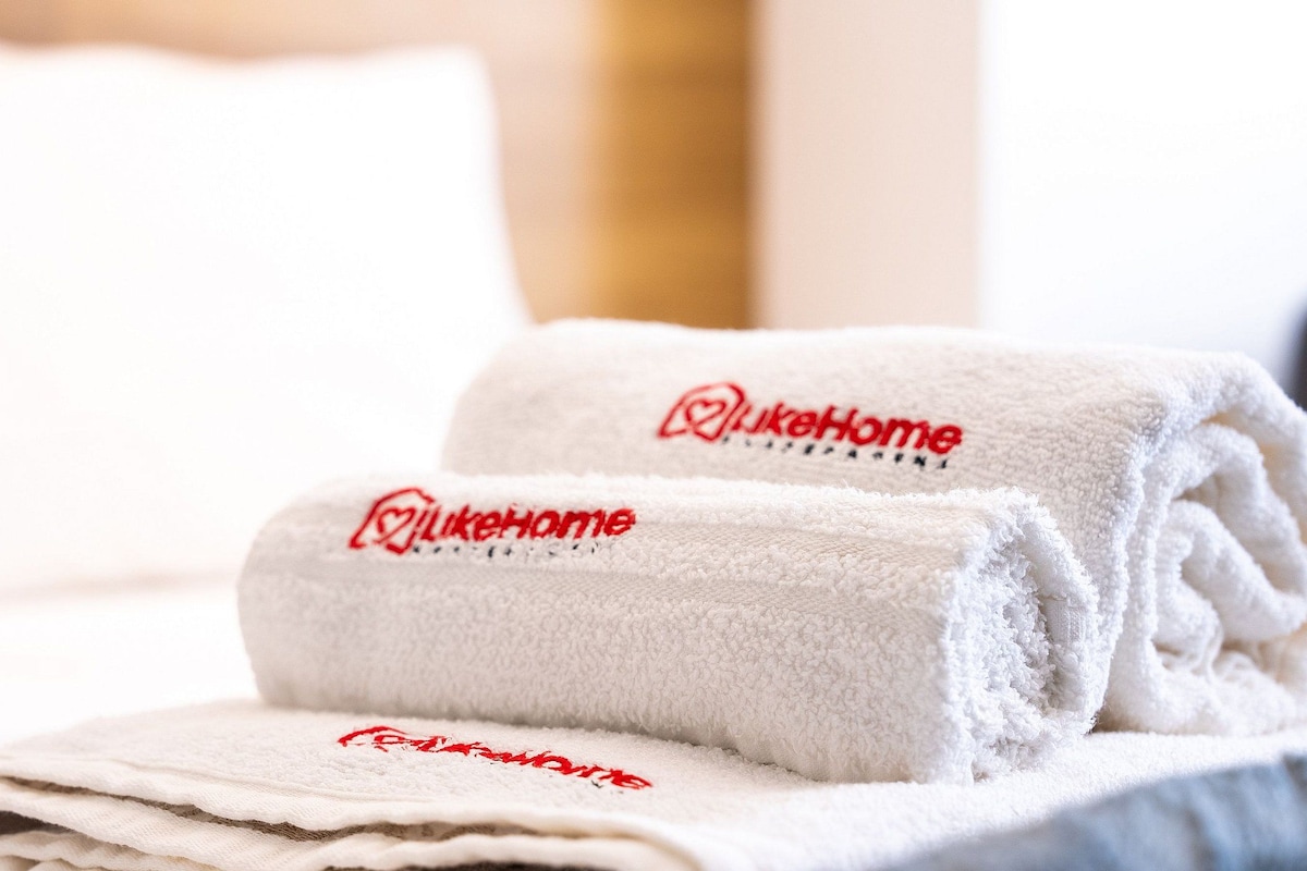 Two neatly folded white towels with 'LukeHome' embroidered in red, placed on a bed.
