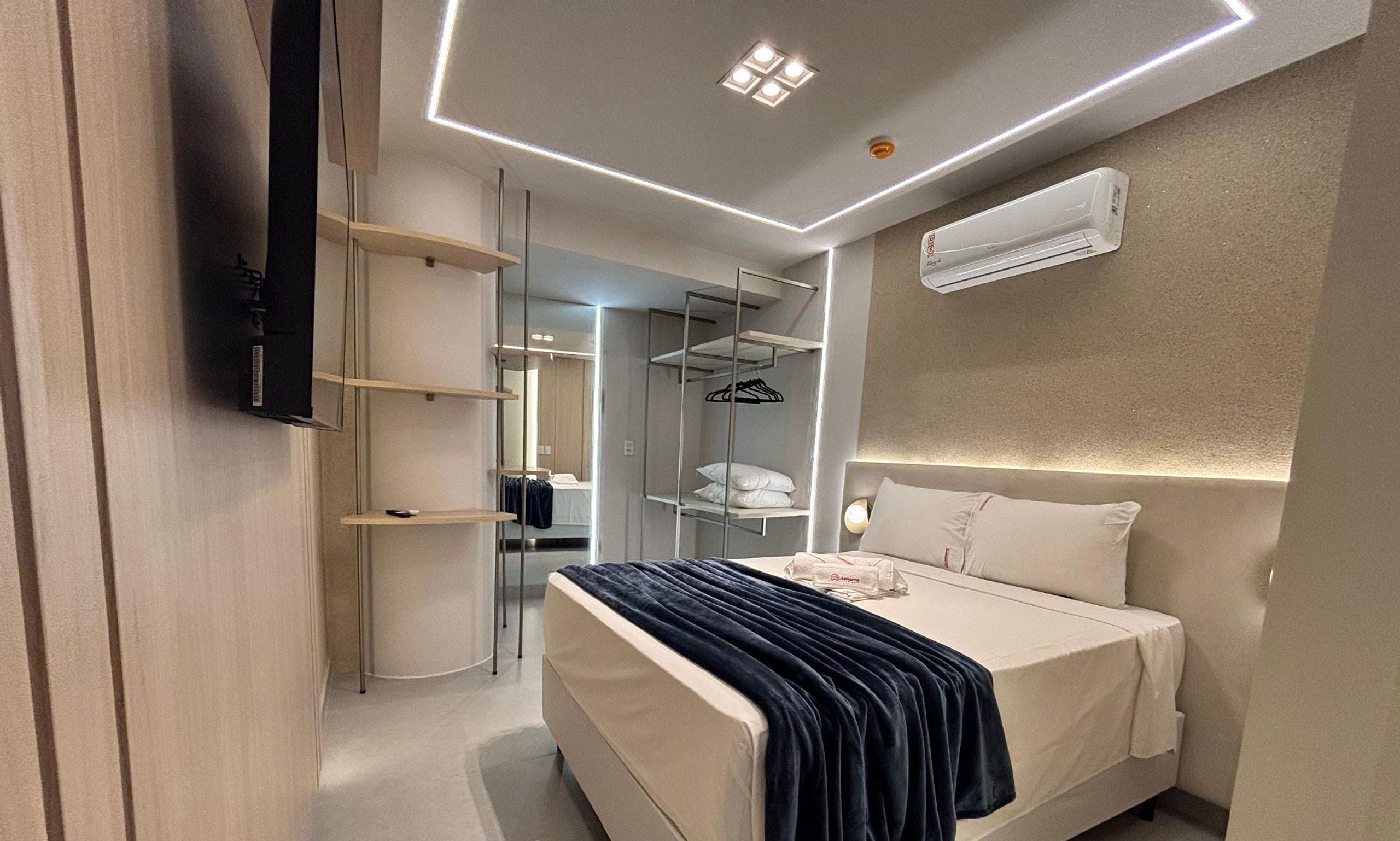 Une chambre d’hôtel moderne équipée d’un lit, d’une table de chevet et d’un téléviseur intégré. On y trouve également une armoire avec un miroir et des étagères, ainsi qu’un climatiseur fixé au mur.