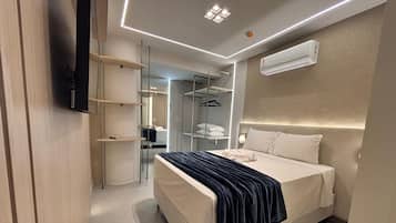 Une chambre d’hôtel moderne équipée d’un lit, d’une table de chevet et d’un téléviseur intégré. On y trouve également une armoire avec un miroir et des étagères, ainsi qu’un climatiseur fixé au mur.