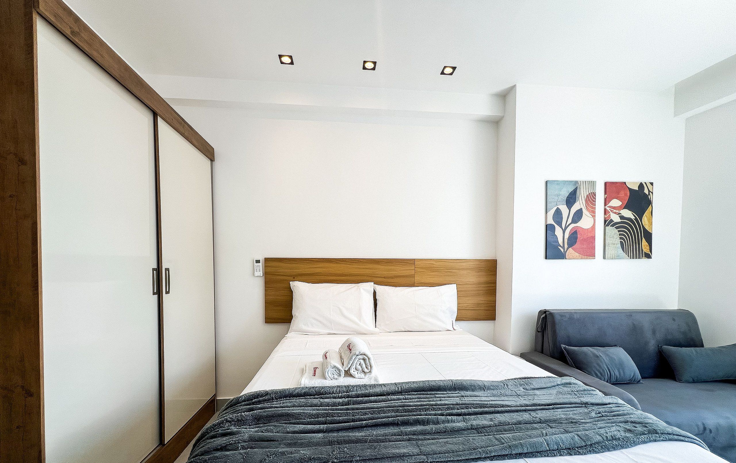 A hotel room with a bed, a wardrobe, a bedside table, a remote control, a grey sofa, and two wall-mounted artworks.