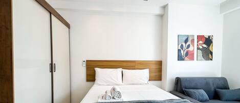 Une chambre d’hôtel comprenant un lit, une armoire, une table de chevet, une télécommande, un canapé gris et deux œuvres d’art accrochées au mur.