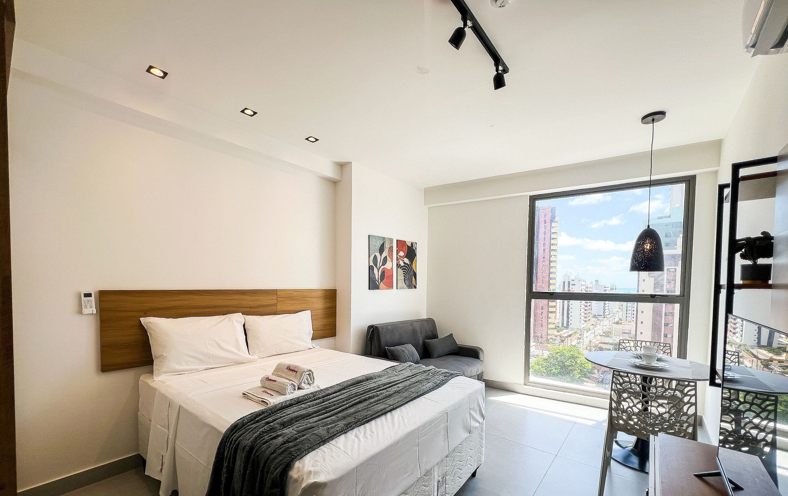 A hotel room with a bed, bedside table, chair, and a large window with a city view.