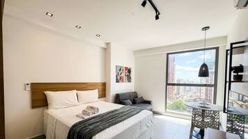 Habitación de hotel con cama, mesita de noche, silla y un amplio ventanal con vista a la ciudad.